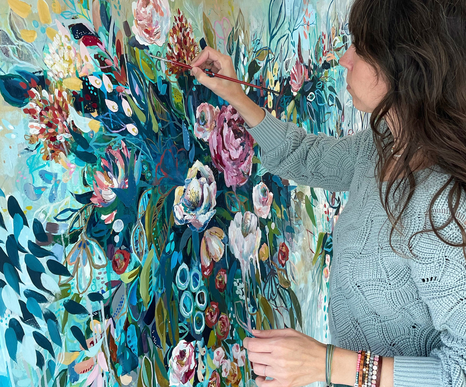 Artist Sunni Mockingbird painting flowers on large stretched canvas in teal, aqua, blue and green leaves with white and pink flowers.